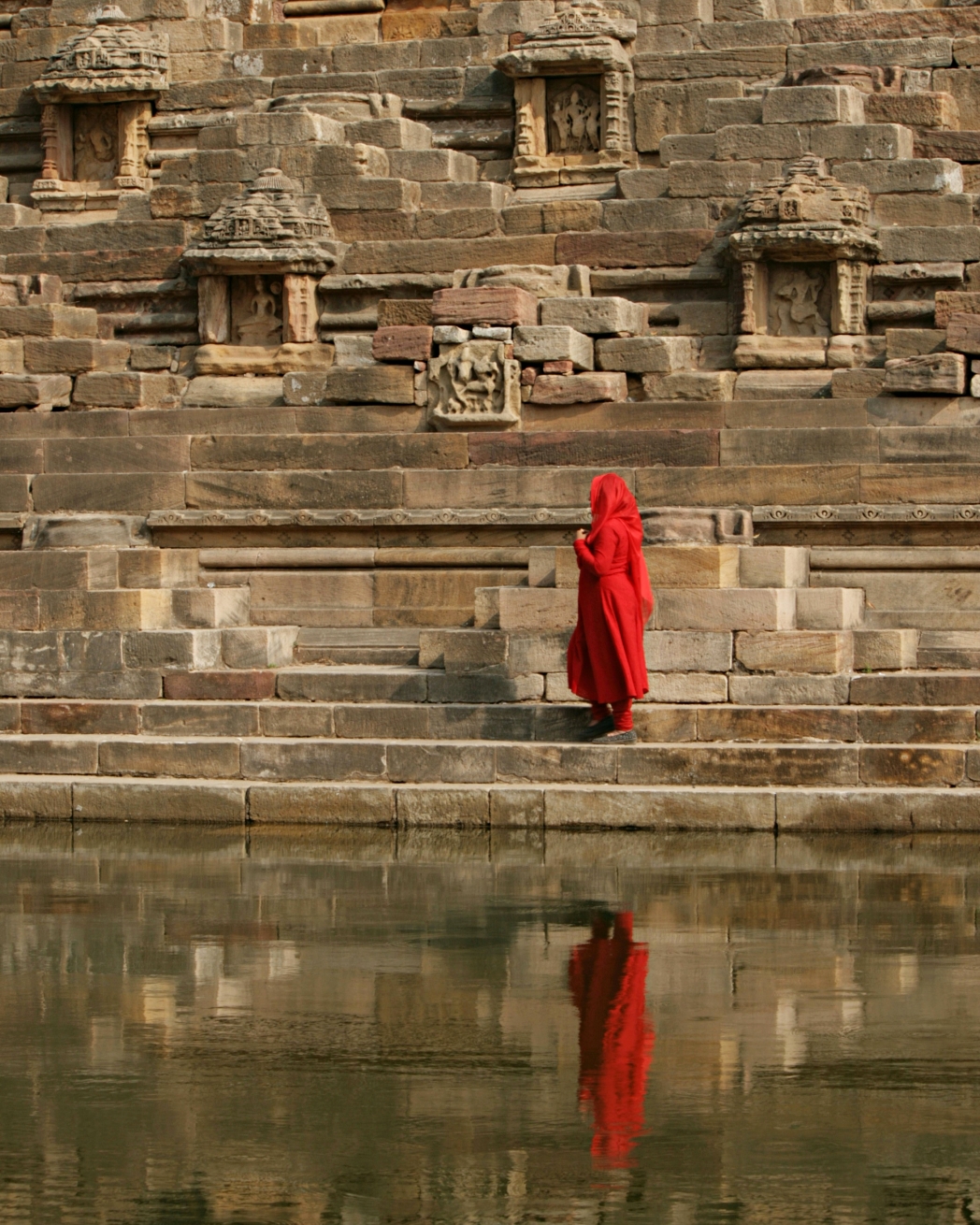 Person in roter Kleidung an einem antiken indischen Stufenbrunnen mit Steinmauern und Wasserspiegelung